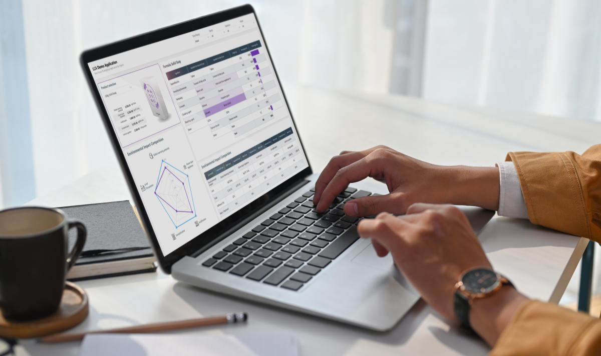 Hands using a laptop showing an LCA analytics dashboard with charts and tables on the screen, with a coffee cup on the desk.